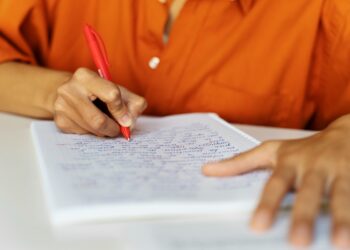 Pessoa escrevendo com caneta vermelha em caderno quadriculado durante prova ou estudo