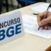 Pessoa preenchendo gabarito de concurso do IBGE, com placa visível ao fundo.