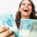 Mulher ri de felicidade enquanto exibe várias cédulas de R$ 100 com a mão estendida