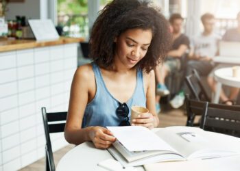 Jovem sorrindo enquanto lê material de estudo em cafeteria com livro aberto e bebida na mão representando preparação para concurso de nível médio