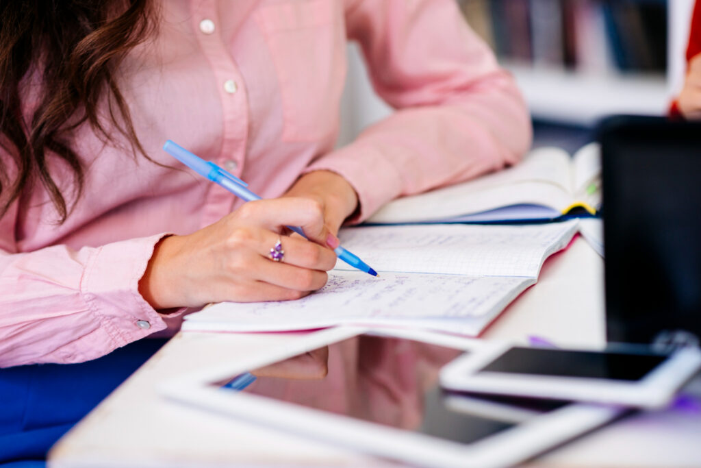 Pare de errar português em concursos! Essas 7 dicas resolvem isso 2 Mulher de camisa rosa anota conteúdos em caderno aberto durante sessão de estudos para concurso público