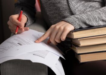 Candidata anotando com lápis vermelho em documentos apoiados sobre pilha de livros durante preparação para o concurso de professor da UFMG com salário de 13,2 mil reais