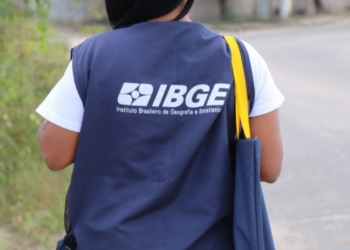 Recenseador do IBGE caminha em rua residencial com colete e bolsa oficiais