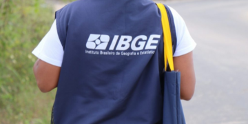 Recenseador do IBGE caminha em rua residencial com colete e bolsa oficiais