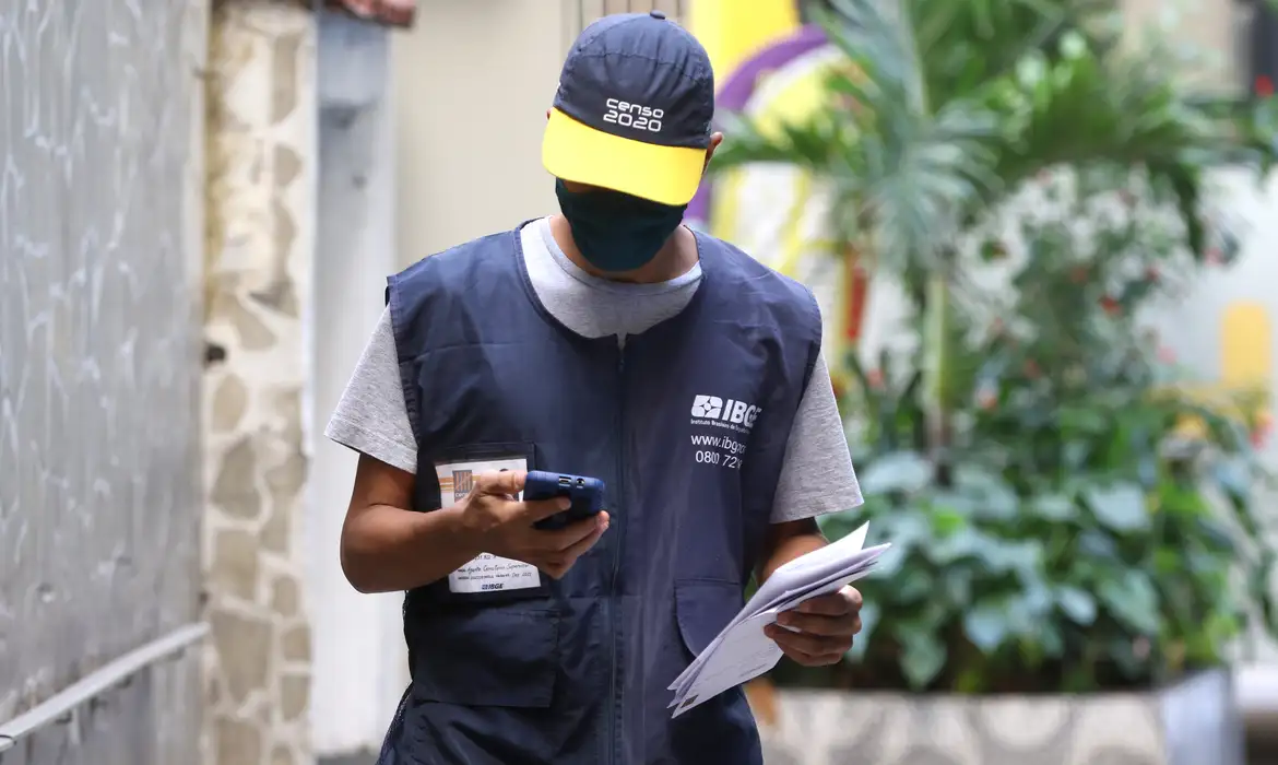 Atenção! IBGE confirma editais de concurso para o 1º semestre do ano 2 Agente do IBGE utilizando celular e documentos durante coleta de dados, ilustrando expectativa pela publicação dos editais do novo concurso ainda no primeiro semestre.