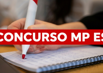 Mão feminina escrevendo com caneta vermelha em um caderno espiral ao lado de notebook