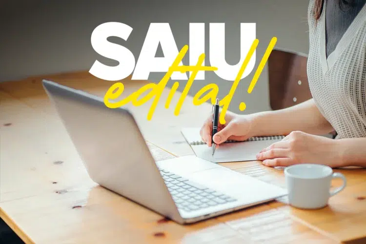 Mulher escreve em caderno ao lado de notebook aberto sobre mesa de madeira