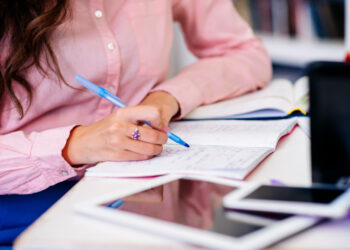 Mão feminina com camisa rosa fazendo anotações em caderno aberto com caneta azul sobre mesa com tablet e smartphone ao lado