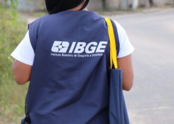 Recenseadora do IBGE caminha com colete azul e bolsa nas ruas de bairro residencial