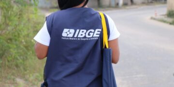 Recenseadora do IBGE caminha com colete azul e bolsa nas ruas de bairro residencial