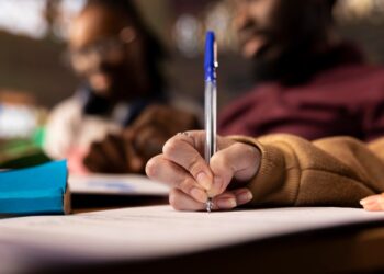 Candidato escrevendo com caneta azul em folha de prova em ambiente de sala de aula representando o concurso da UFMA com remuneração de até R$ 4,9 mil