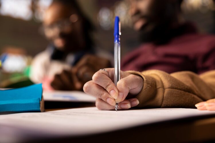 Saiu edital! UFMA abre concurso com 59 vagas e remuneração de até R$ 4,9 mil 1 Candidato escrevendo com caneta azul em folha de prova em ambiente de sala de aula representando o concurso da UFMA com remuneração de até R$ 4,9 mil