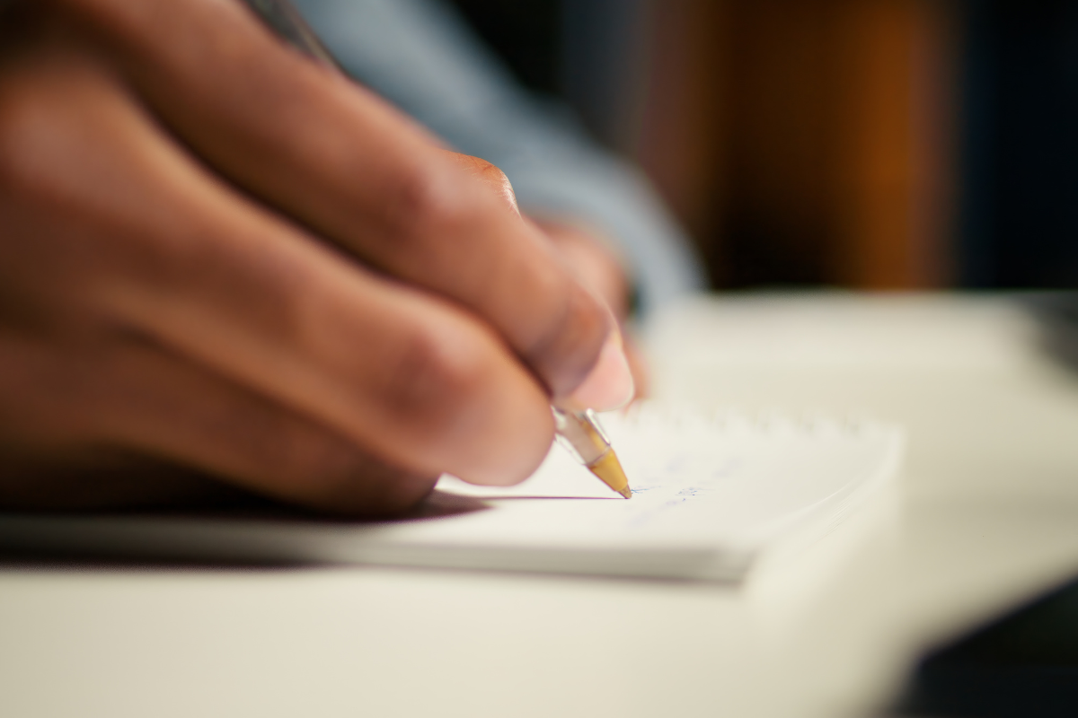 SAIU EDITAL! Concurso com vagas para Nutricionista, Psicólogo, Farmacêutico e outros cargos está com inscrições abertas - Salários de até R$ 9,4 mil 2 Mão segurando caneta escreve anotações em folha branca com foco seletivo