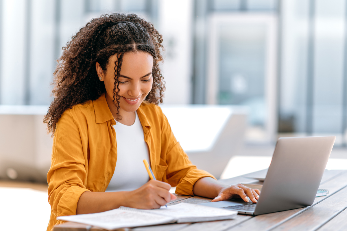Reprovou no concurso? Respira! Falhar faz parte do caminho e não significa que acabou 2 Mulher com camisa amarela anota em caderno enquanto utiliza notebook em mesa iluminada