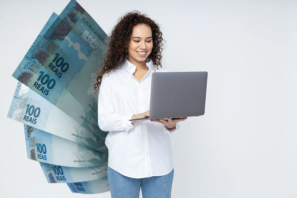 Concursos: Quais são os cargos públicos com os maiores salários? 2 Mulher de cabelos cacheados usando notebook em frente a cédulas de 100 reais