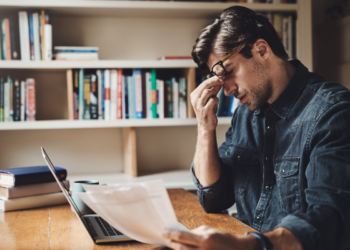 Homem sentado à mesa observa papéis com expressão preocupada diante de laptop