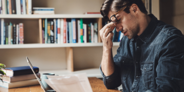 Homem sentado à mesa observa papéis com expressão preocupada diante de laptop