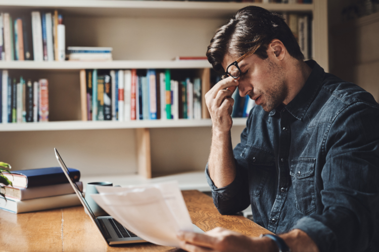 Reprovou no concurso? Respira! Falhar faz parte do caminho e não significa que acabou 1 Homem sentado à mesa observa papéis com expressão preocupada diante de laptop