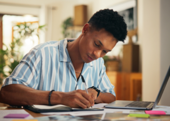Homem escreve em caderno ao lado de notebook em mesa com papéis coloridos