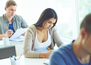 Jovem mulher morena de cabelo liso concentrada escrevendo com lápis em sala de aula clara com outros estudantes desfocados ao redor