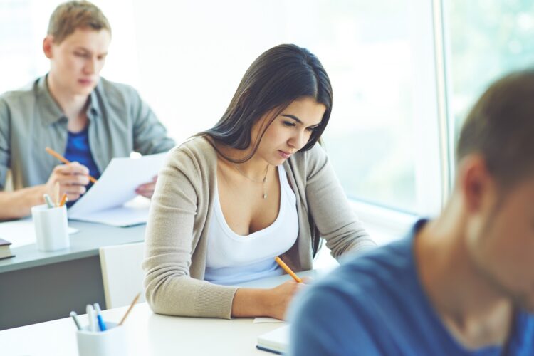 2 novos editais oferecem mais de 300 vagas e salários de até R$ 11,9 mil 1 Jovem mulher morena de cabelo liso concentrada escrevendo com lápis em sala de aula clara com outros estudantes desfocados ao redor