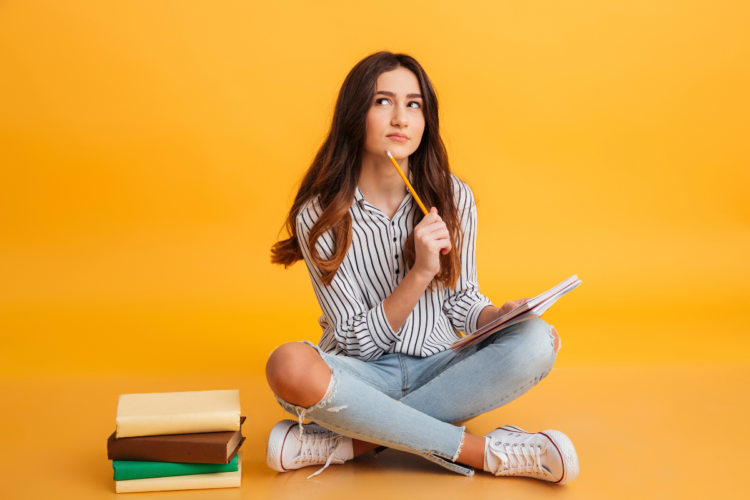 Quer se preparar para concursos antes do edital? Veja cinco dicas que fazem a diferença 1 Jovem sentada no chão, segura caderno e lápis, olha pensativa ao lado de livros empilhados