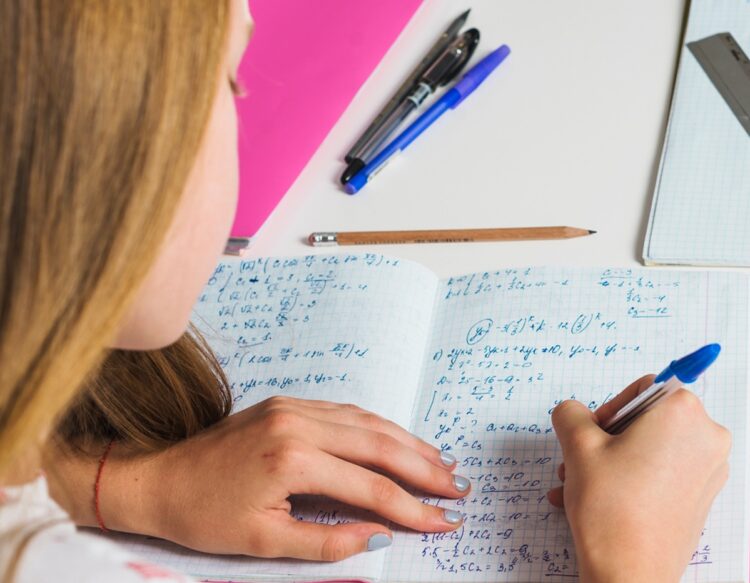 Como estudar Matemática para concursos públicos: 5 dicas que funcionam 1 Mulher jovem de cabelo cacheado concentrada escrevendo em livro aberto com caneta verde em mesa de estudo com copo e caderno ao lado