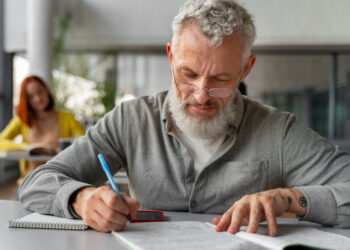 Homem de cabelos grisalhos e óculos estudando concentrado com caneta azul e caderno aberto em sala de aula representando a preparação para concursos após os 40 anos