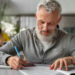 Homem de cabelos grisalhos e óculos estudando concentrado com caneta azul e caderno aberto em sala de aula representando a preparação para concursos após os 40 anos
