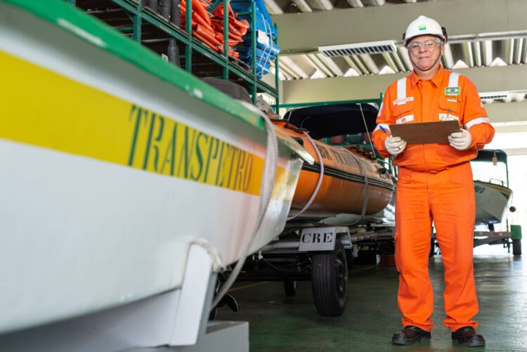 Concurso Transpetro: edital está em elaboração e previsão de publicação já anima candidatos 2 Profissional com uniforme laranja e prancheta inspeciona barcos em galpão Transpetro