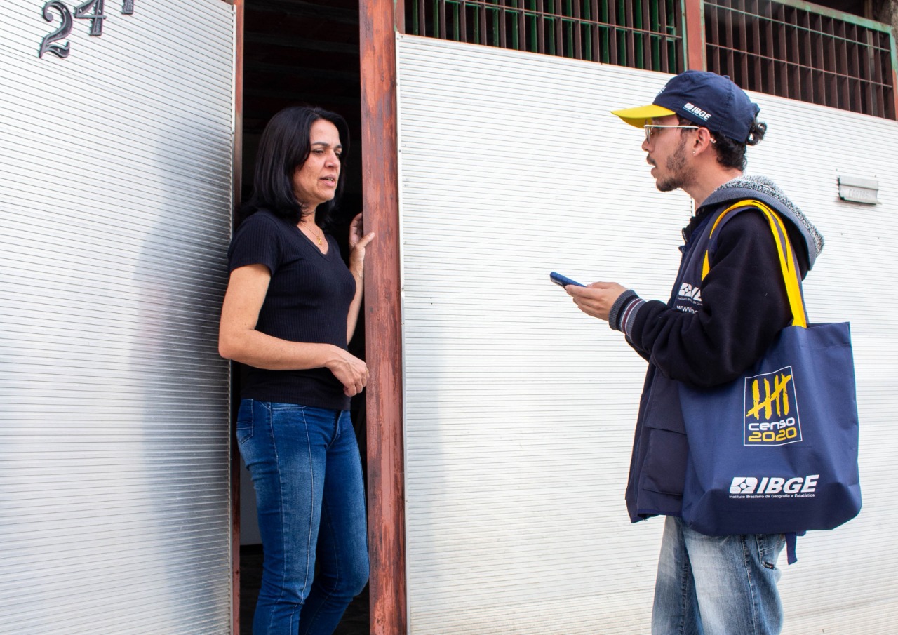 Qual o melhor cargo no concurso temporário do IBGE para você? Veja como descobrir 2 Recenseador uniformizado do IBGE entrevista mulher na porta de casa usando dispositivo móvel