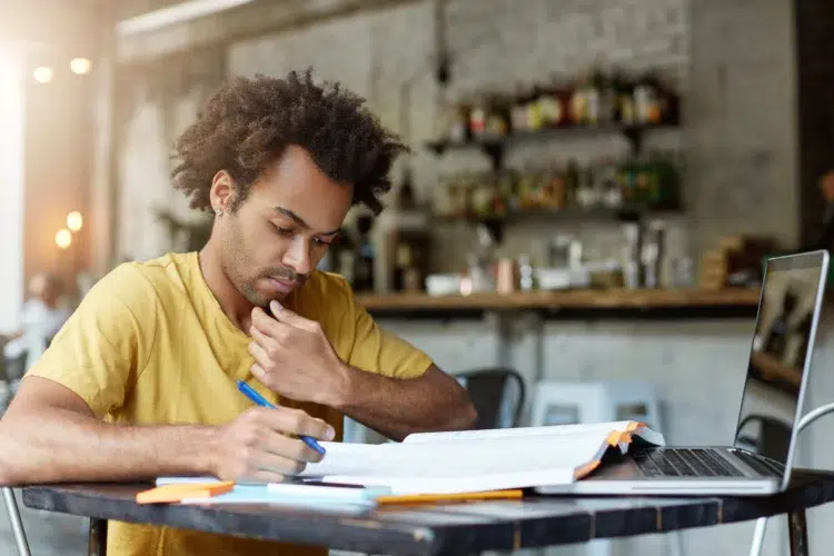 Como usar a IA para estudar para concursos: 5 prompts que aumentam a produtividade 2 Jovem negro de cabelo crespo e camiseta amarela estuda com livro, caderno e laptop em mesa de café.