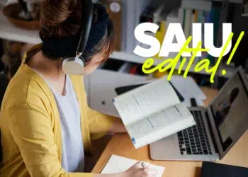 Mulher de cardigan amarelo e fone estuda em mesa com notebook, livro aberto e texto “SAIU edital!” na tela.