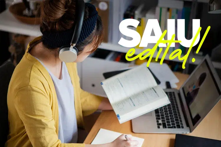 Concurso em São Gonçalo do Rio Abaixo - MG tem vagas para Agente Comunitário de Saúde 1 Mulher de cardigan amarelo e fone estuda em mesa com notebook, livro aberto e texto “SAIU edital!” na tela.