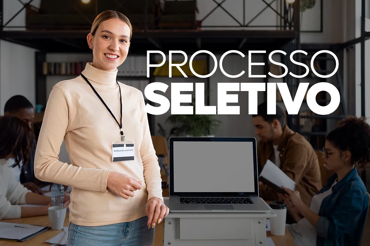 Prefeitura de Três Marias - MG lança processo seletivo com remuneração de até R$ 19,5 mil 2 Mulher jovem sorridente com crachá em pé ao lado de um notebook aberto, com o texto "PROCESSO SELETIVO" em destaque e profissionais trabalhando ao fundo.