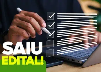 Mão segurando caneta stylus branca diante de notebook com checklist digital e texto “Saiu Edital!” em destaque.