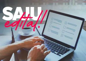 Mãos digitando em laptop com checklist na tela, xícara de café ao lado e texto "SAIU edital!" em destaque.