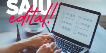 Mãos digitando em laptop com checklist na tela, xícara de café ao lado e texto "SAIU edital!" em destaque.