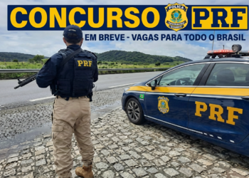 Policial rodoviário federal armado ao lado de viatura PRF em rodovia pavimentada com paisagem montanhosa