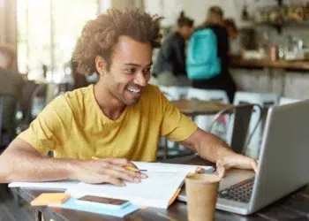 Homem jovem de cabelo crespo e camiseta amarela sorri enquanto estuda com notebook e papéis em cafeteria.