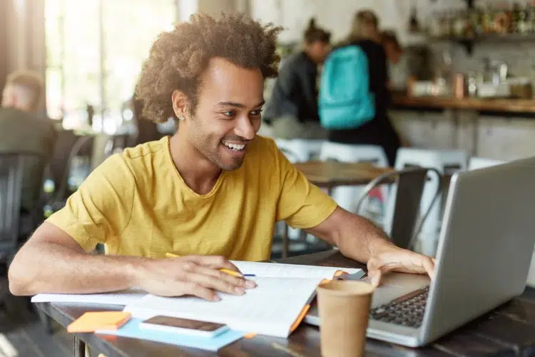 Homem jovem de cabelo crespo e camiseta amarela sorri enquanto estuda com notebook e papéis em cafeteria.