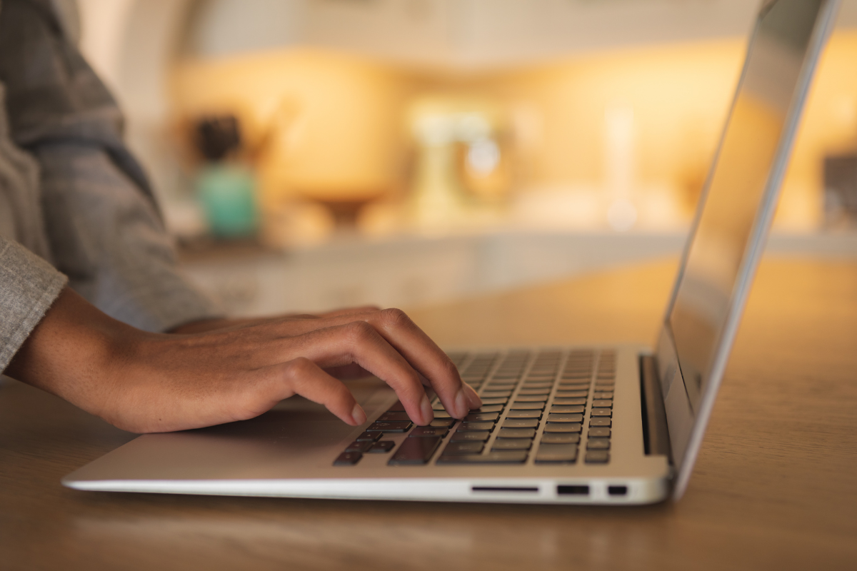 Assembleia Legislativa de Roraima abre concursos com salários de até R$ 39,5 mil 2 Mão feminina digitando no teclado de notebook prateado em mesa de madeira com iluminação ambiente quente ao fundo.