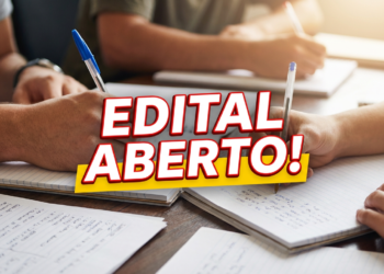 Estudantes escrevem em cadernos sobre mesa de madeira com destaque para letreiro "Edital Aberto!" em vermelho e amarelo.