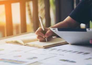 Pessoa segurando caneta prateada escreve em caderno amarelo sobre mesa com gráficos coloridos e tablet.