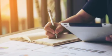 Pessoa segurando caneta prateada escreve em caderno amarelo sobre mesa com gráficos coloridos e tablet.
